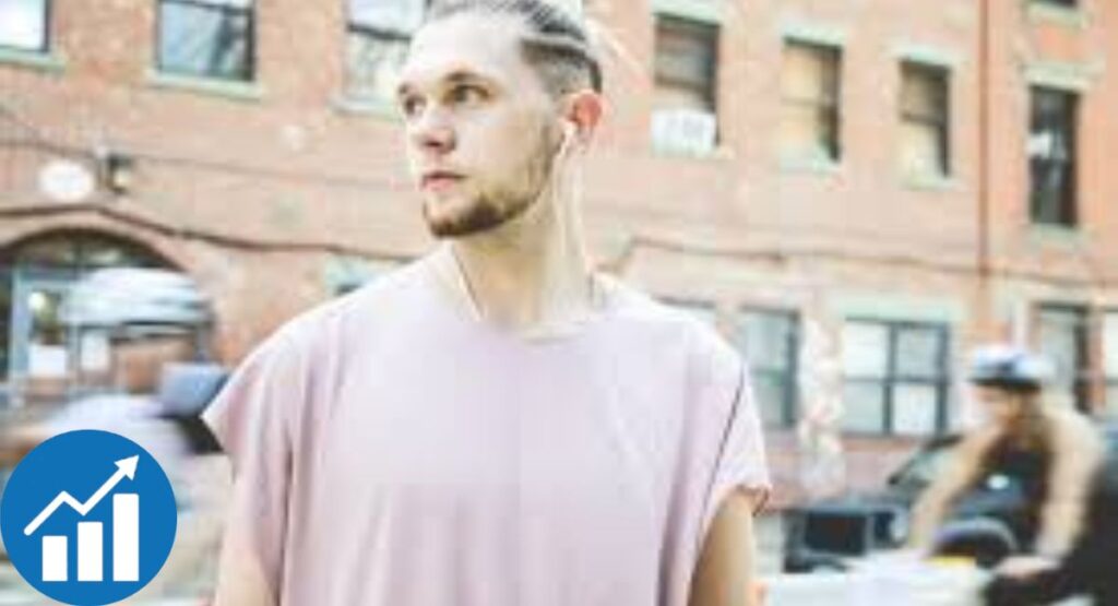 Brandon Kee wearing a light pink sleeveless shirt, standing outdoors in an urban setting with earphones in.