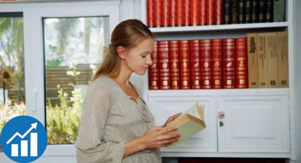 Ivi Rein reading a book in a modern study with a white bookshelf, red hardcover volumes, and garden view.